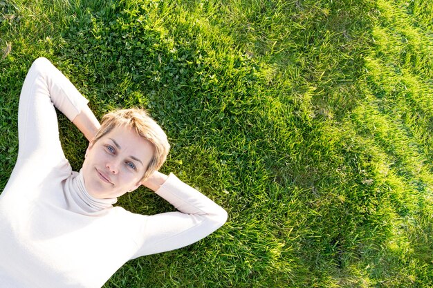 Woman with short hair in white top resting on meadow green grass on sunny spring warm day in park