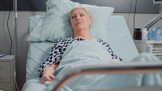 Woman with sickness receiving treatment with IV drip bag, sitting in hospital ward bed. Retired patient connected to oxygen tube for respiratory problem, trying to help with healthcare