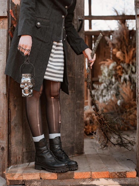 Woman with skull candle holder and broom while standing on doorstep of wooden house during halloween