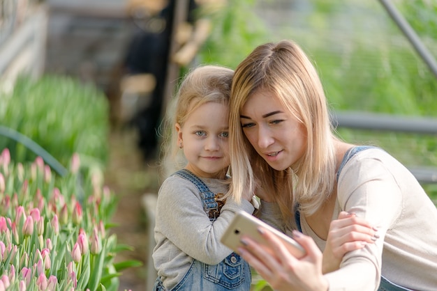 A woman with a small daughter take a selfie in a greenhouse with flowers in spring