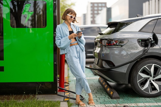 スマートフォンを持った女性が電気自動車の充電を待っています