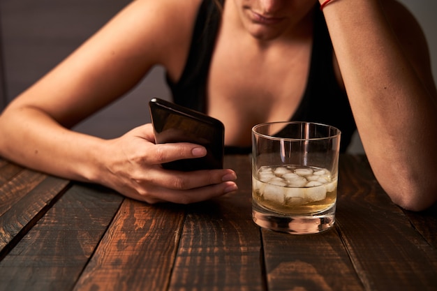 Woman with a smartphone and a glass of alcohol at a bar. Concept of alcoholism.