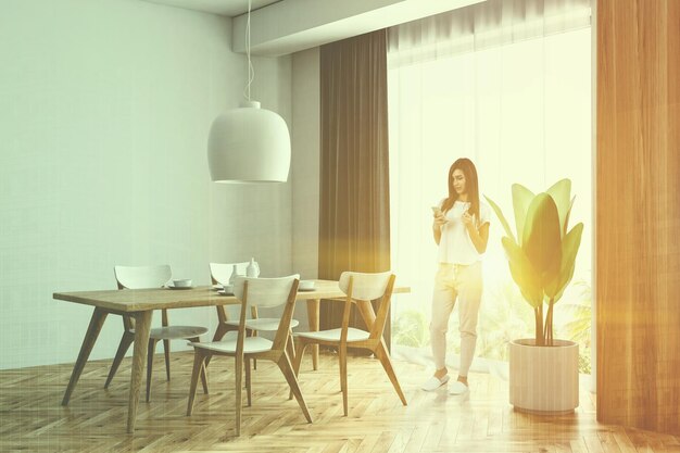 Woman with a smartphone in a white and wooden dining room corner with a wooden floor, a wooden table with chairs and a loft window. 3d rendering mock up toned image double exposure