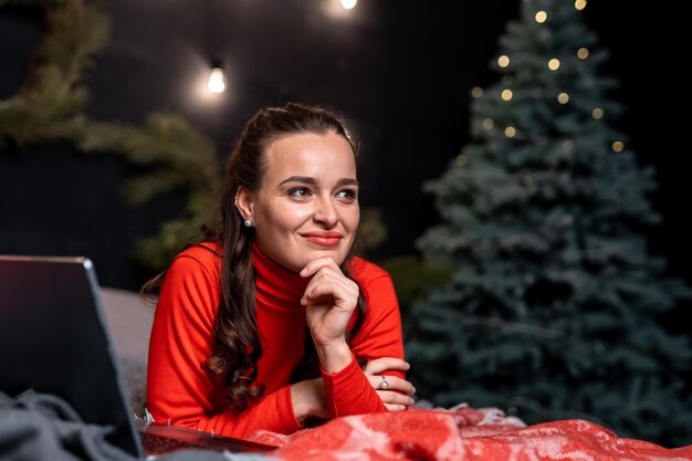 Woman with a smile on the red blanket A lady with a dreamy smile lying on the plaid with a laptop by her side Lying girl with a notebook near the Christmas tree