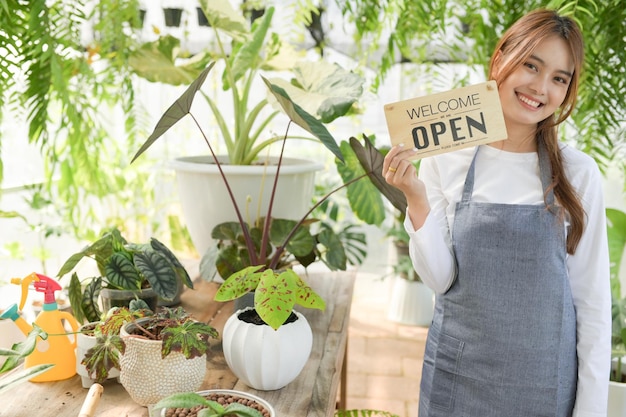 Photo a woman with a smiling face in an apron stands holding a welcome open sign in the green house