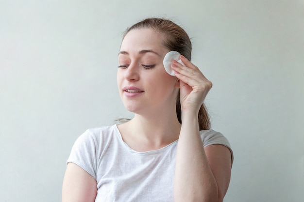 Woman with soft healthy skin removing make up with cotton pad isolated on white wall