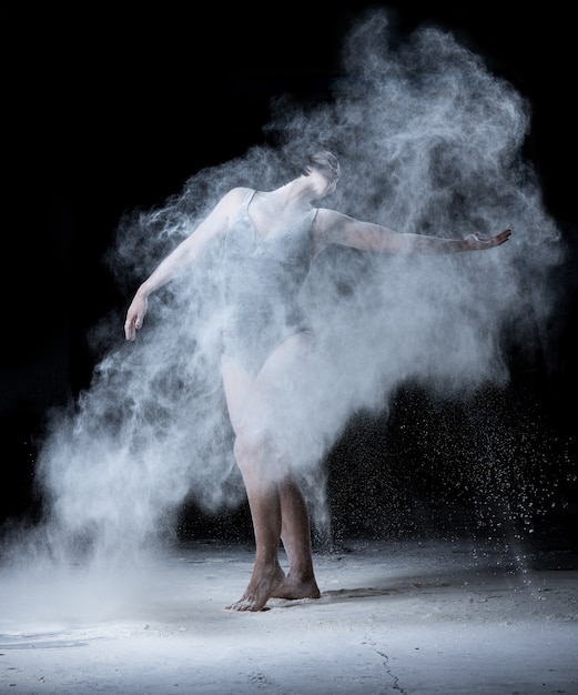Woman with a sports figure dancing in a cloud of white scattered flour on a black background, the dancer is dressed in a black bodysuit