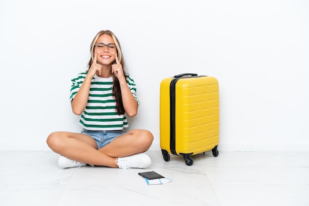 Woman with suitcase sitting on the floor smiling with a happy and pleasant expression