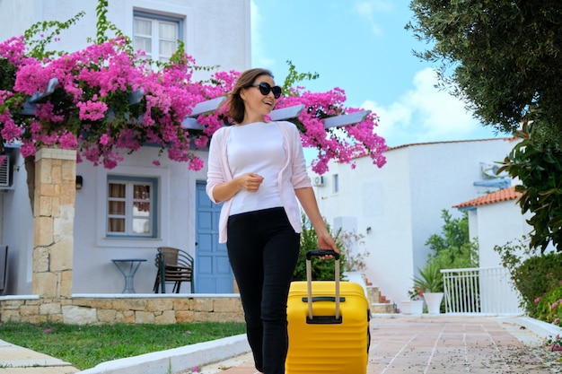 美しいピンクの花の造園と海の絵のように美しいリゾートスパホテルの領域を屋外で歩くスーツケースを持つ女性。旅行、休暇、レジャー、週末、人々