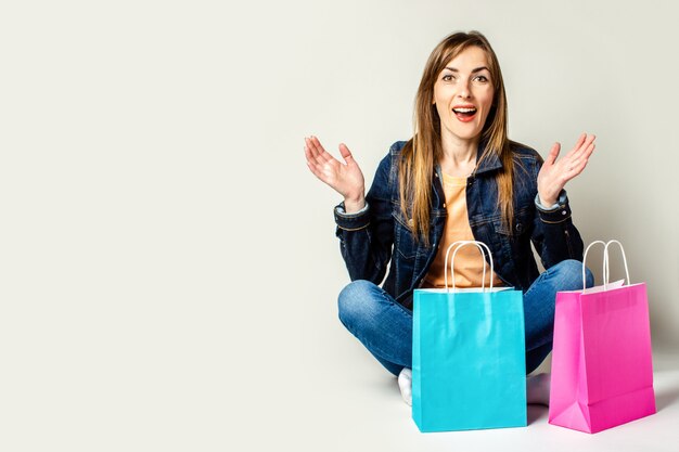 Woman with a surprised face in a denim jacket holds shopping bags while sitting on the floor on a light space. Banner. Concept shopping.