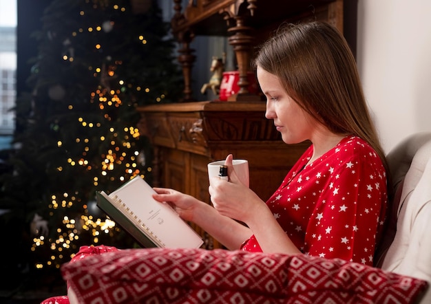 Woman with tea cup in hands sitting in chair at cozy home with fir tree on christmas holidays