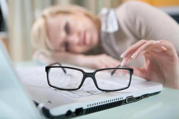 Фото Женщина с усталыми глазами в очках лежит на столе рядом с ноутбуком, сосредоточенным на очках