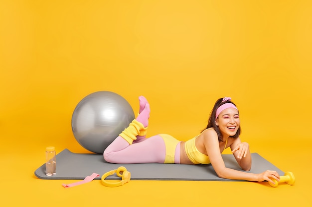 woman with tired glad expression lies on fitness mat surrounded by sport equipment dressed in sportswear 