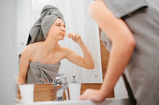 Woman with a towel on her head brushes her teeth while looking in the bathroom mirror