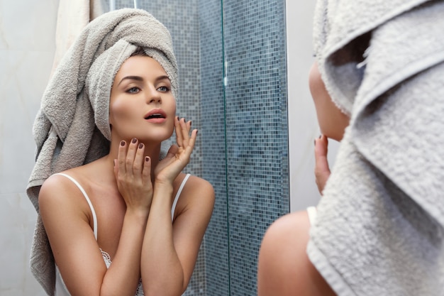 Woman with a towel on her head, looking in the mirror and applying make up