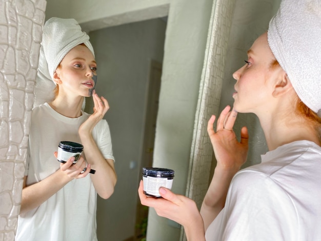 Woman with a towel on her head puts a clay mask on her face at home