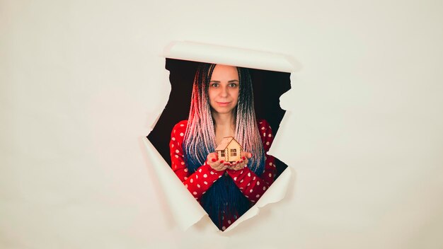 woman with toy house looking through hole in paper female with afro braids and miniature wooden house looking thro torn white paper background and showing concept of real estate