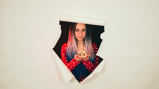 Woman with toy house looking through hole in paper Female with Afro braids and miniature wooden house looking thro torn white paper background and showing concept of real estate