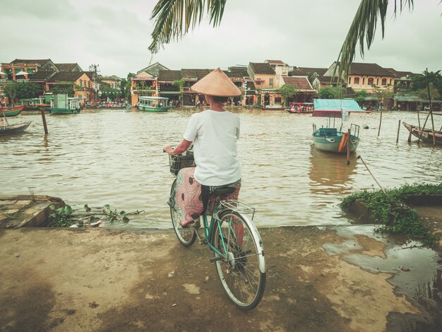 Woman with traditional vietnamese hat cycling on the river bank at Hoi.