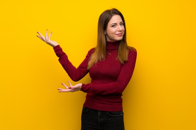 Donna con dolcevita sul muro giallo che estende le mani a lato per invitare a venire