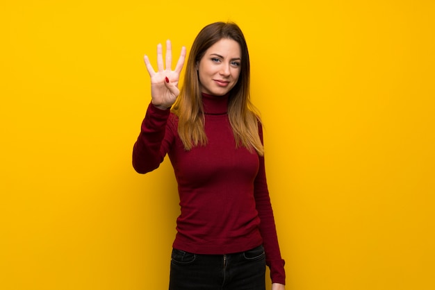 Donna con dolcevita sul muro giallo felice e contando quattro con le dita