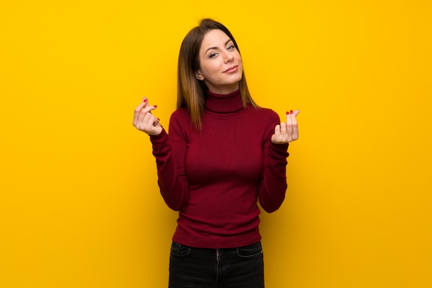 Woman with turtleneck over yellow wall making money gesture