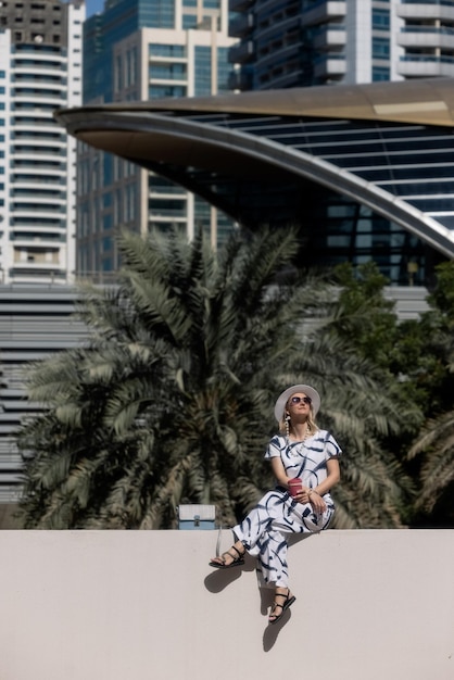 Woman with a white hat and coffe is sitting on a balcony in front of the Dubai metro station