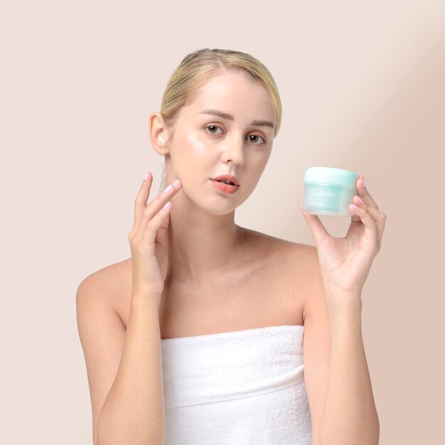 A woman with a white towel on her head holds a jar of face cream.