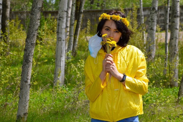 Woman with wreath of dandelions and medical mask smelling bouquet of dandelions