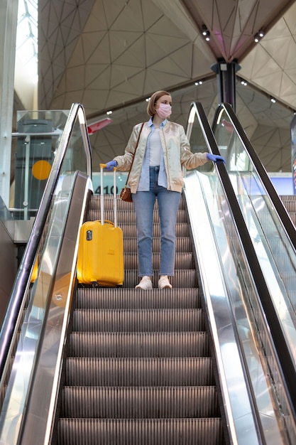 Woman with yellow luggage stands on escalator at almost empty airport terminal due to coronavirus pandemic