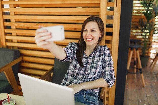 Donna in legno all'aperto street summer coffee shop seduto con computer pc portatile, facendo selfie girato sul telefono cellulare, rilassante durante il tempo libero. ufficio mobile