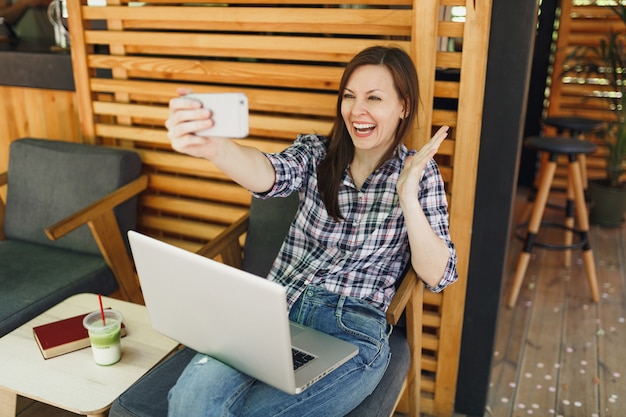 Donna in legno all'aperto street summer coffee shop seduto con computer pc portatile, facendo selfie girato sul telefono cellulare, rilassante durante il tempo libero. ufficio mobile