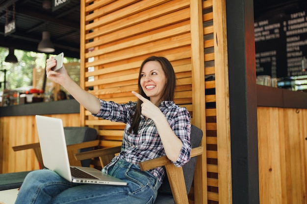 Woman in wooden outdoors street summer coffee shop sitting with laptop pc computer, doing selfie shot on mobile phone, relaxing during free time. Mobile Office