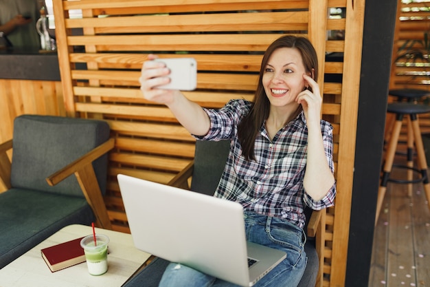 Woman in wooden outdoors street summer coffee shop sitting with laptop pc computer, doing selfie shot on mobile phone