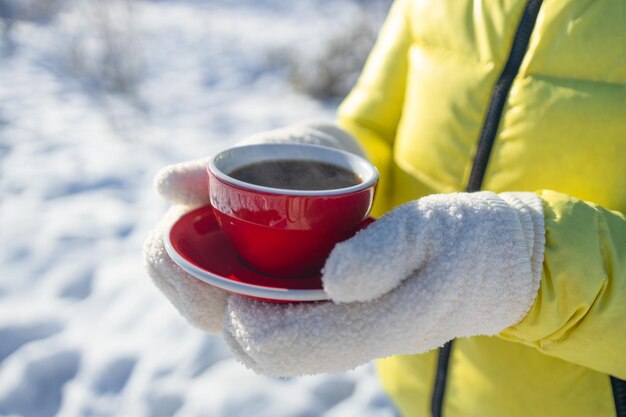 羊毛の手袋をかぶった女性が冬の街の外で赤いカップのコーヒーを保持し温かい飲み物を飲んでいます