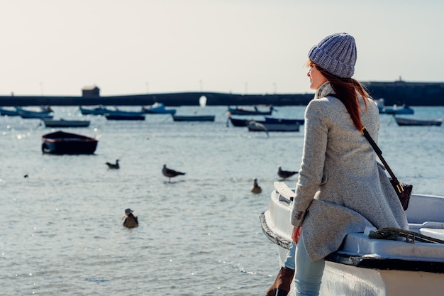 Женщина в шерстяной шапке опирается на рыбацкую лодку и смотрит на море