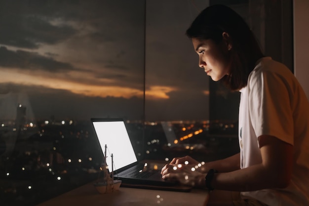 Женщина работает с ноутбуком у окна квартиры с вечерним видом на город