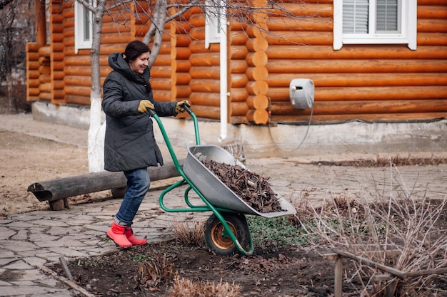 Woman worker in field clothes applying mulching procedure spreading old dried grass in barrow Seasonal working in garden Organic fertilizer for best harvest homemade compost humus Eco harvest