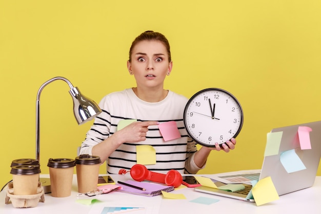 Photo woman worker pointing finger at big wall clock in her hands sitting at workplace working late hours