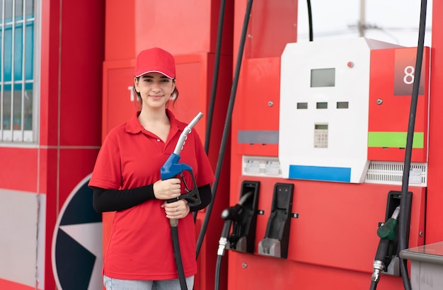 Woman worker in red uniform holding fuel pump nozzle ready to service at gas station