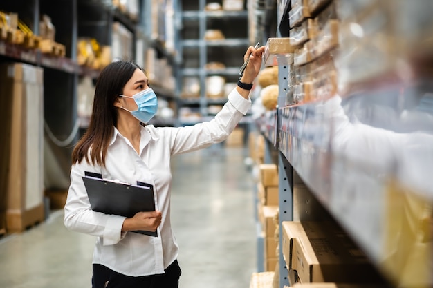 Lavoratrice con maschera medica che tiene appunti e controlla l'inventario nel magazzino durante la pandemia di coronavirus