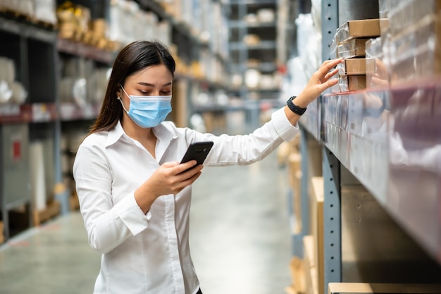 Woman worker with medical mask using smartphone to check inventory in the warehouse during coronavirus pandemic