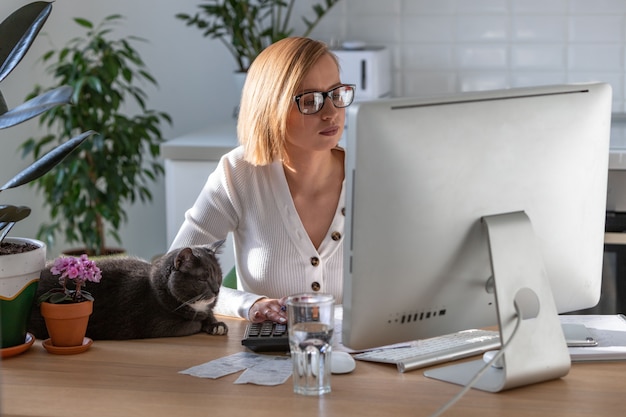 Woman working on computer from home office during self-isolation period, cat sleeps nearby on the table