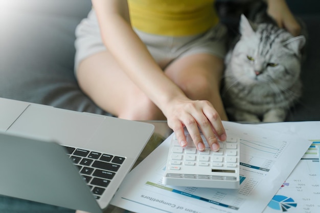 사진 소파 에 고양이 와 함께 앉아 있는 집 에서 일 하는 여자 가 계산기 를 사용 한다