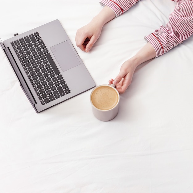 Женщина работая от дома с пижамой компьтер-книжки нося в кровати. Она пьет кофе и работает на кровати. Вид сверху и копирование пространства.