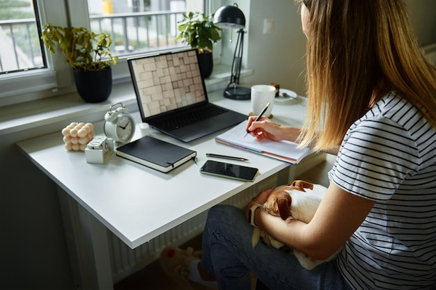 Woman working at home office use laptop and hold dog on hands