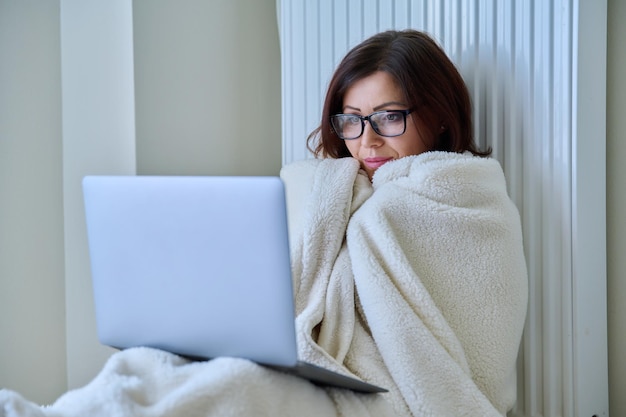 Woman working at home with laptop warming with blanket and heating radiator