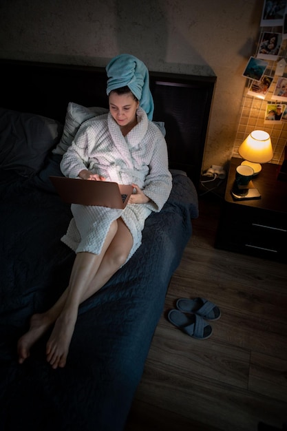 Woman working on laptop at home laying in bed wet head covered with towel freelance concept