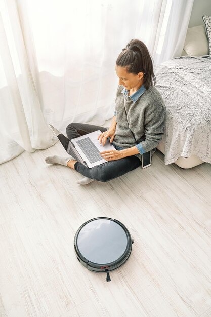 Woman working on laptop while robotic vacuum cleaner cleaning\
floor