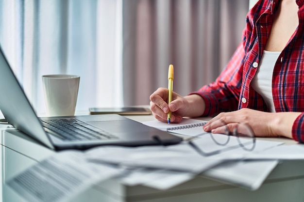 Woman working online on laptop and writing down data information in notebook. Female during studying remotely at home